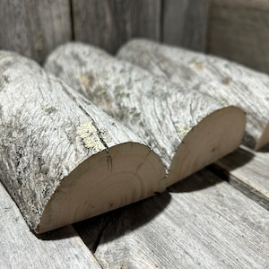 May include: Three weathered, gray-toned wooden logs with visible bark and lichen. The logs are cut in half lengthwise, revealing the light-colored wood grain. The logs are arranged on a weathered wooden surface, creating a rustic aesthetic.