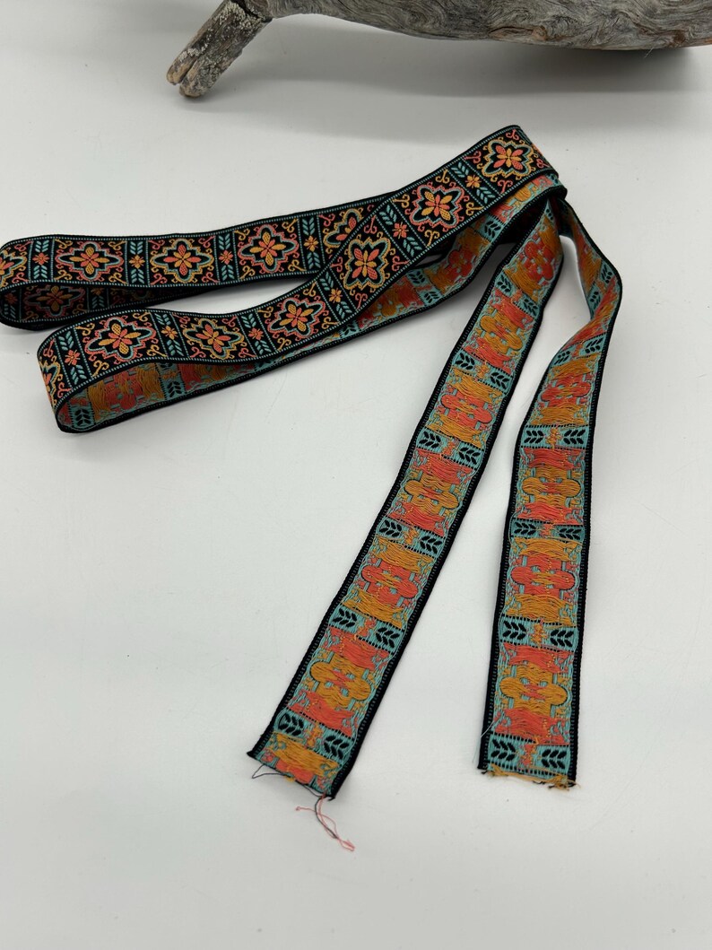 May include: A length of decorative ribbon with a black background and a repeating pattern of orange and teal floral and geometric designs. The ribbon is likely used for crafting or embellishment.