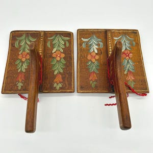 May include: Two wooden hand fans with a floral design. The fans have a dark brown background with green leaves, orange flowers, and red accents. The handles are made of wood and are attached with red string.