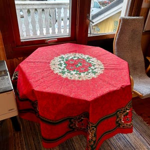 Op de afbeelding: Een rode, achthoekige tafelkleed met een centraal bloemmotief met rode poinsettia's, groene bladeren en gouden accenten. De rand heeft een complementair ontwerp. Het tafelkleed is waarschijnlijk voor een feestdag.