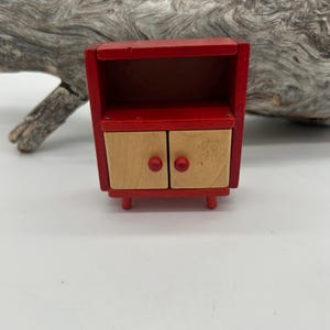 May include: A miniature red wooden cabinet with two natural wood-toned doors and red knobs. The cabinet has a small shelf and is supported by four short legs. The design is simple and the cabinet appears to be handmade.
