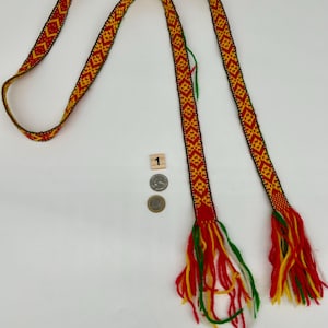 May include: A long, woven belt featuring a repeating geometric diamond pattern in red, yellow, and green. The ends of the belt are finished with loose, multi-colored yarn tassels in red, yellow, green, and white. A small wooden block with the number 1 and two coins are placed near the center for scale.