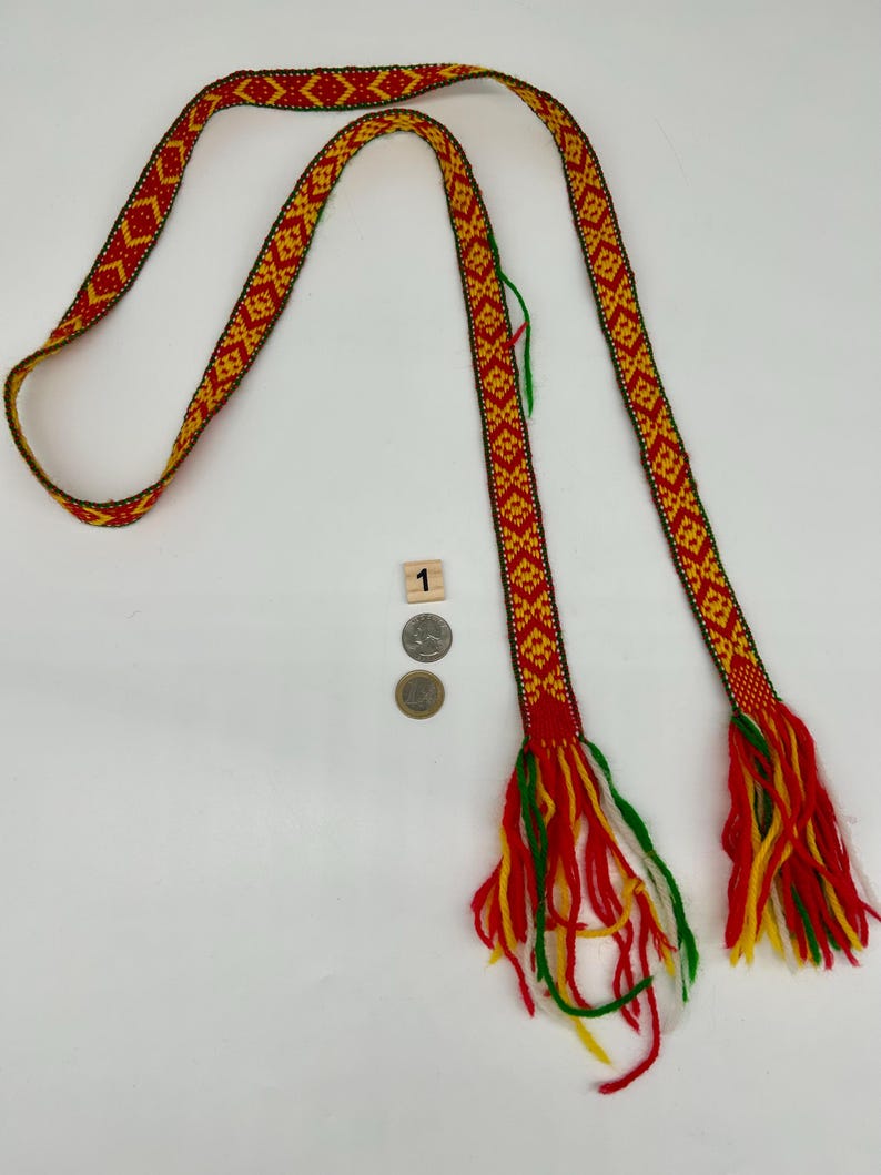 May include: A long, woven belt featuring a repeating geometric diamond pattern in red, yellow, and green. The ends of the belt are finished with loose, multi-colored yarn tassels in red, yellow, green, and white. A small wooden block with the number 1 and two coins are placed near the center for scale.