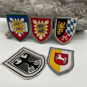 May include: A collection of five embroidered patches with heraldic designs. The patches feature various shield shapes with emblems like lions, horses, and eagles. The patches have a variety of colors, including red, yellow, blue, black, and white.