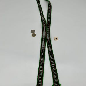 May include: A long, narrow woven textile band featuring a repeating geometric pattern in green, white, and pink. The strip is folded in half, displaying fringed ends. Two circular coins and a small square tile with the number 5 are visible.