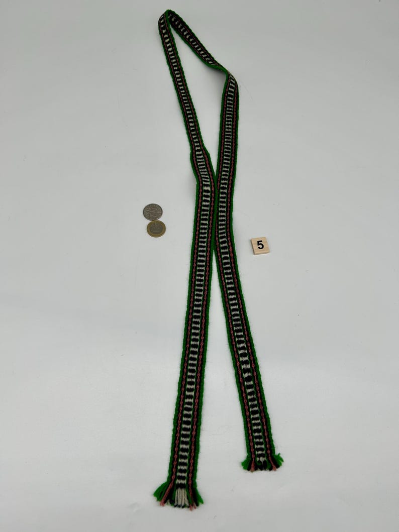 May include: A long, narrow woven textile band featuring a repeating geometric pattern in green, white, and pink. The strip is folded in half, displaying fringed ends. Two circular coins and a small square tile with the number 5 are visible.