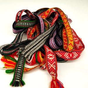 May include: A collection of woven fabric ribbons featuring intricate geometric patterns in vibrant colors including red, yellow, green, black, and white. The ribbons are tangled together, with some ends showing loose, multi-colored yarn tassels.