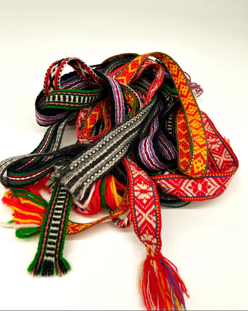 May include: A collection of woven fabric ribbons featuring intricate geometric patterns in vibrant colors including red, yellow, green, black, and white. The ribbons are tangled together, with some ends showing loose, multi-colored yarn tassels.