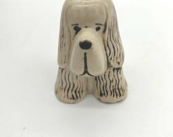 May include: A ceramic dog figurine in a seated position. The dog has a light beige body with dark brown detailing on the face and fur. The figurine has a glossy finish and a classic, vintage aesthetic.