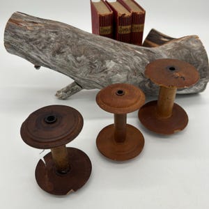 Può includere: Tre antiche bobine di legno con finitura marrone scuro, disposte davanti a un tronco di legno trasportato dall'acqua. Piccoli libri rossi sullo sfondo. Le bobine hanno un design classico, con una parte superiore e inferiore circolari collegate da uno stelo centrale.