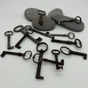 May include: A collection of antique keys in various sizes and designs, with dark brown and silver tones. Some keys are resting on gray stones, while others are scattered on a white surface. The keys have ornate details and different handle shapes.