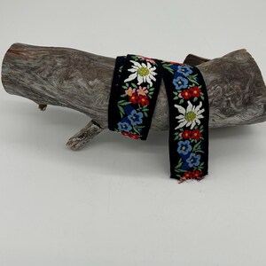 May include: Black embroidered ribbon with a floral pattern featuring white, red, blue, and yellow flowers. The ribbon is displayed on a piece of weathered wood against a white background. The ribbon's design includes detailed stitching.