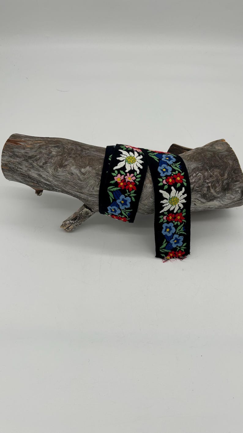 May include: Black embroidered ribbon with a floral pattern featuring white, red, blue, and yellow flowers. The ribbon is displayed on a piece of weathered wood against a white background. The ribbon's design includes detailed stitching.