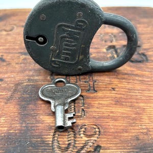 Puede incluir: Un candado antiguo de color verde oscuro con su llave. El candado tiene la palabra "Yale" grabada en su superficie. La llave es plateada y también tiene la palabra "Yale". El candado y la llave están sobre una superficie de madera.