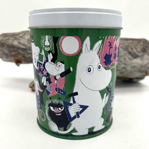 May include: A green metal tin with a white lid, featuring cartoon characters in various poses. The tin's design includes white, pink, and black illustrations on a green background. The characters are from the Moomin series.