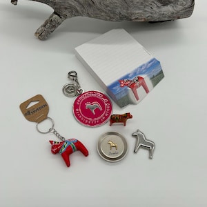 Può includere: Una collezione di souvenir svedesi a tema cavallo Dala. Include un portachiavi a cavallo Dala rosso, un ciondolo circolare rosa, un blocco note con un'immagine di cavallo Dala e una statuina di cavallo.