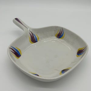 Peut inclure: Plat de service en céramique blanche avec une poignée. Le plat est de forme carrée aux coins arrondis et décoré d'un motif floral jaune, bleu et violet. La poignée est blanche avec un accent violet.
