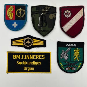 May include: A collection of embroidered patches in various shapes and colors. Designs include a shield with a cross, a shield with a letter "B", a shield with a diagonal stripe, a yellow bar with a plane, and a rectangular patch with text.