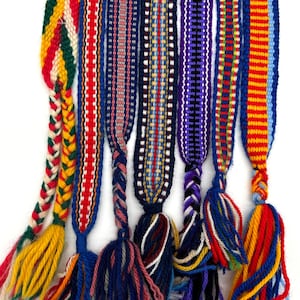 May include: A selection of vibrantly coloured woven belts, each with a braided section and a tassel. The belts display diverse patterns and colour schemes, including red, blue, yellow, and green. The tassels are made of yarn, adding a decorative touch.
