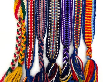 May include: A collection of colorful woven belts with braided and tasseled ends. The belts feature various patterns and color combinations, including red, blue, yellow, and green. The tassels are made of yarn and add a decorative element to the belts.