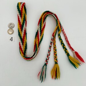 Pu&ograve; includere: Una cintura tessuta con un motivo colorato di rosso, verde, giallo e bianco. La cintura ha una sezione lunga e piatta e tre nappe intrecciate all'estremit&agrave;. Il numero 4 &egrave; visibile accanto ad alcune monete.