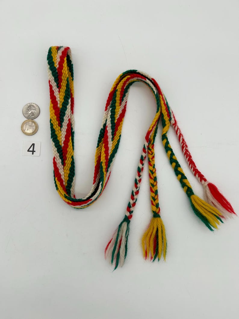 Pu&ograve; includere: Una cintura tessuta con un motivo colorato di rosso, verde, giallo e bianco. La cintura ha una sezione lunga e piatta e tre nappe intrecciate all'estremit&agrave;. Il numero 4 &egrave; visibile accanto ad alcune monete.