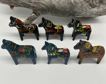 May include: Six hand-painted wooden Dala horses in various colors, including black, blue, and brown. Each horse features a unique floral design in red, yellow, and green. The horses are displayed on a white surface with a piece of driftwood in the background.