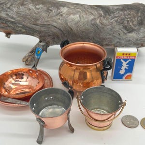 Könnte beinhalten: Eine Sammlung von Miniatur-Kupferkochgeschirr, darunter ein Topf, eine Pfanne und ein Eimer. Die Kupferstücke haben eine glänzende Oberfläche und werden von einer kleinen Streichholzschachtel mit blau-weißen Grafiken begleitet. Ein Stück Treibholz befindet sich im Hintergrund.