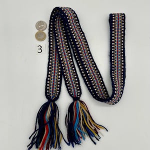 Pu&ograve; includere: Una cintura tessile intrecciata con sfondo blu navy e un motivo a righe rosse, bianche e gialle. La cintura ha nappe alle due estremit&agrave; con un mix di fili blu, rossi, gialli e color crema. Il numero "3" &egrave; visibile.