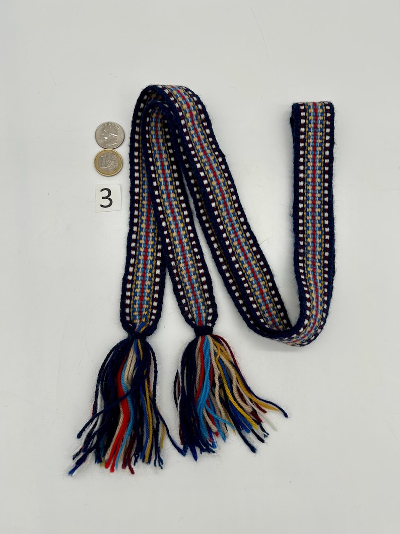 Pu&ograve; includere: Una cintura tessile intrecciata con sfondo blu navy e un motivo a righe rosse, bianche e gialle. La cintura ha nappe alle due estremit&agrave; con un mix di fili blu, rossi, gialli e color crema. Il numero "3" &egrave; visibile.