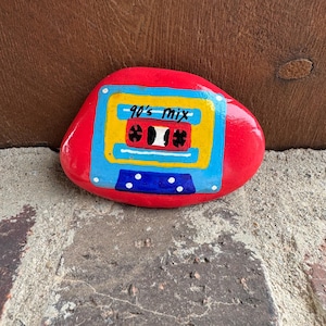 May include: A hand-painted red rock with a retro cassette tape design. The cassette is yellow, blue, and black, with the text "90's mix". A unique, decorative item.