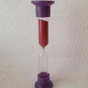 Könnte beinhalten: Eine Sanduhr mit einem durchsichtigen Glaskern und einem lila Kunststoffdeckel und -sockel. Die Sanduhr ist mit rotem Sand gefüllt. Die oberen und unteren Teile sind zylindrisch und die Basis ist kreisförmig.