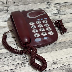 May include: A burgundy rotary phone with a black coiled cord. The phone has a keypad with numbers 1 through 9, a star, a zero, and a pound sign. The phone has a red dial button.