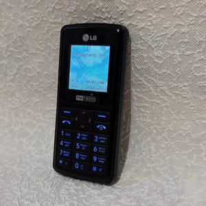 Op de afbeelding: Zwarte LG mobiele telefoon met een blauw verlicht scherm met tekst en een hemelsblauwe achtergrond. De telefoon heeft een toetsenbord met blauwe cijfers en letters. Het scherm toont de tekst "Незарегистр, STH" en "FM radio".