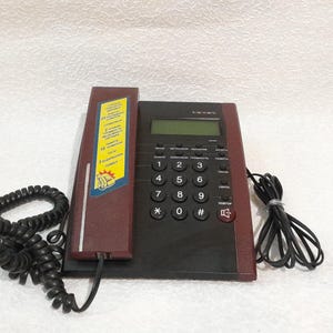 Op de afbeelding: Een vintage telefoon met snoer in bordeauxrood en zwart. De telefoon heeft een numeriek toetsenbord, een klein beeldscherm en een opgerold handset snoer. Aan de zijkant zit een sticker met tekst.