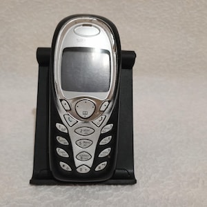 Puede incluir: Un teléfono móvil negro y plateado con teclado y pantalla, apoyado en un soporte negro. El teléfono tiene un diseño curvo con un borde plateado y una pantalla. El teclado presenta números y símbolos blancos.