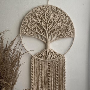 Könnte beinhalten: Beige Makramee-Wandbehang mit einem Baumdesign in einem kreisförmigen Rahmen. Der Baum hat detaillierte Äste und Wurzeln mit langen Fransen darunter. Das Kunstwerk besteht aus geknotetem, naturfarbenem Kordel, wodurch eine strukturierte, böhmische Ästhetik entsteht.