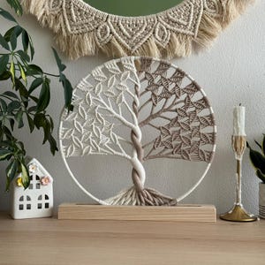 Op de afbeelding: Een ronde macramé wandhanger met een boomontwerp in een tweekleurig schema van crèmekleurig en lichtbruin koord. De boom is opgebouwd met ingewikkelde knooppatronen en is gemonteerd op een rechthoekige, lichtgekleurde houten voet.