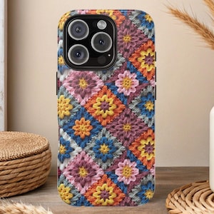 Puede incluir: Una funda de teléfono colorida con un diseño de cuadrados de la abuela. La funda presenta un patrón de patchwork de cuadrados rosas, naranjas, amarillos, azules y morados. La funda del teléfono está sobre una superficie de madera.