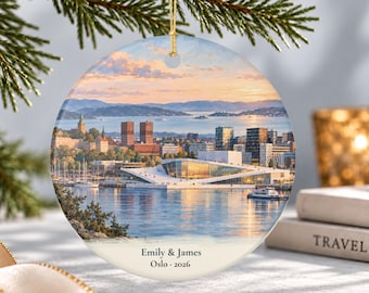 May include: A round ceramic ornament featuring a sunset view of the Oslo waterfront, including the Opera House and city skyline. The bottom section is cream-colored with the printed text "Emily & James" and "Oslo · 2026" in black font.