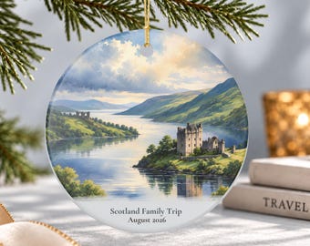 May include: A round ceramic ornament featuring a landscape painting of a Scottish castle on a lake with green hills and a cloudy sky. The text "Scotland Family Trip" and "August 2026" is printed in black at the bottom of the white surface.