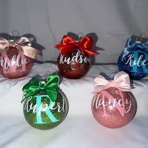 May include: Five personalised Christmas baubles with glitter and bows. The baubles are pink, red, green, blue, and gold. The names on the baubles are Millie, Hudson, Riley, Rupert, and Nancy.
