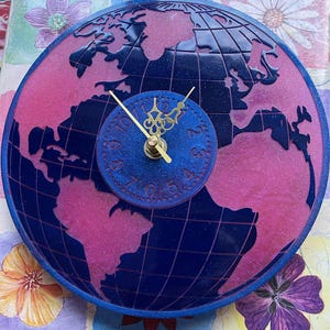 Può includere: Un orologio rotondo decorativo con un design a mappa del mondo. Il quadrante è un cerchio più piccolo con lancette e numeri dorati. Il design del globo presenta continenti rosa e oceani blu.