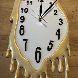 Könnte beinhalten: Eine weiße Wanduhr mit schwarzen Ziffern und Zeigern, die ein einzigartiges Schmelzdesign aufweist. Der Rand der Uhr hat einen goldenen, tropfenden Effekt, der ein surreales und künstlerisches Aussehen erzeugt. Die Uhr befindet sich auf einer Holzoberfläche.