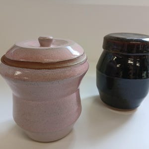 Könnte beinhalten: Zwei Keramikgläser. Eines ist hellrosa mit Deckel und kleinem Knauf. Das andere ist ein dunkelbraunes, zylindrisches Glas mit Deckel. Beide Gläser haben eine glänzende Oberfläche und stehen vor einem weißen Hintergrund.