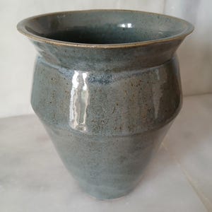 Può includere: Un vaso in ceramica blu-verde fatto a mano con un bordo ampio e leggermente svasato e una superficie strutturata. Il vaso ha un corpo arrotondato che si assottiglia verso la base. L'interno e il bordo hanno una tonalità blu più chiara.