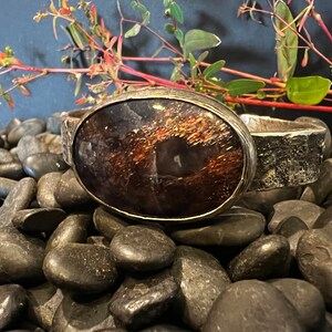 May include: A silver cuff bracelet with a large oval-shaped gemstone set in the center. The gemstone is a deep brown color with iridescent gold and black accents. The bracelet is resting on a bed of smooth black stones.