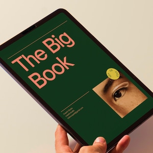 May include: A green tablet with a dark grey border displays a book cover with the title "The Big Book" in a peachy-pink font. The cover also features a close-up of a person's eye with a yellow sticker that says "Off 2021".