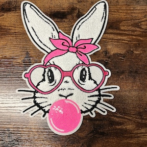 May include: A white bunny patch with a pink bow, heart-shaped glasses, and a bubblegum bubble. The bunny has black whiskers and is set against a wooden background. The patch is likely for clothing or accessories.
