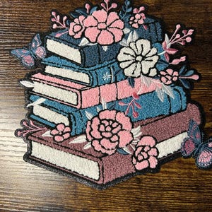 May include: Embroidered patch depicting a stack of books in pink, teal, and burgundy, decorated with pink and white flowers and butterflies. The patch has a black border and a textured finish, ideal for clothing or accessories.